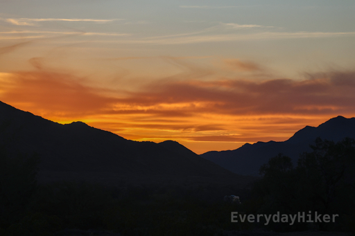 Sunset taken from the Ironwood National Monument BLM land near the Saguaro National Park. Two shadowed mountain ridges may be seen leading from opposite sides of the frame in the distance, while a brilliant orange glow burns above them. Small bands of clouds may be seen in the sky which turns to blue along the upper frame.