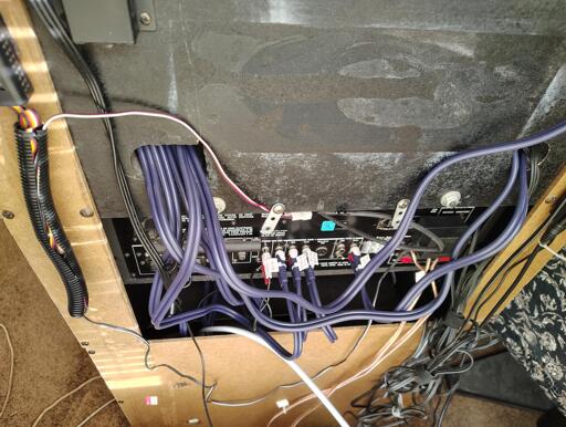 The back side of an old fiberboard stereo cabinet. Cables are draped throughout the scene.  Most of the cables are a bluish purple colour and go into an opening at the top left.  the other ends of the cables are plugged into sockets on the back of a receiver and have small  white tags indicating which device they're associated with.  At the top left, a length of black plastic sheath hides most of the cables dangling from a variable-voltage regulator designed for PC case fans.