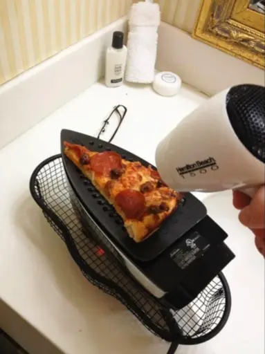 A pizza slice on the underside of a clothing iron with a hair dryer blowing on it